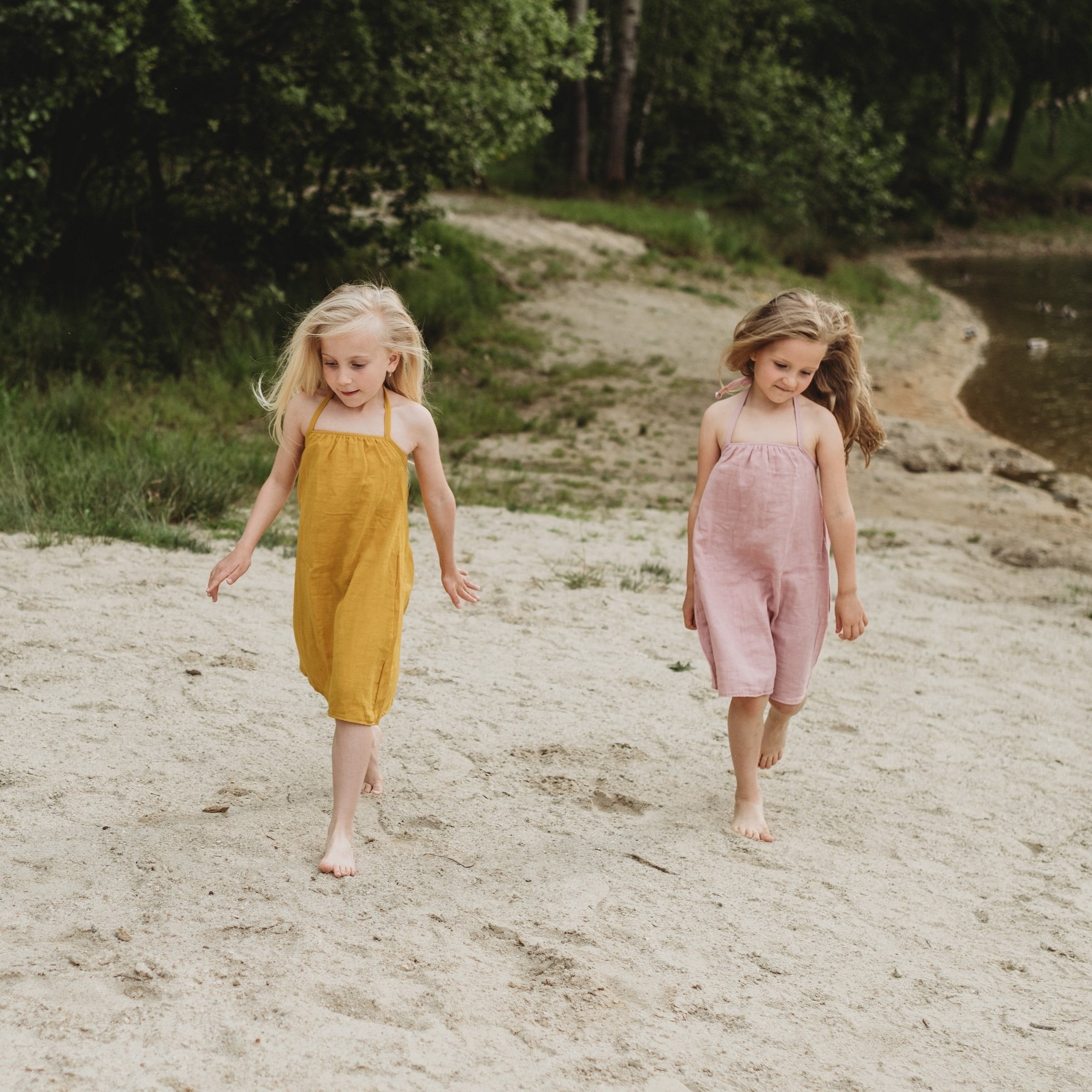 Two girls walking on a beach wearing organic muslin dresses in mustard and pink